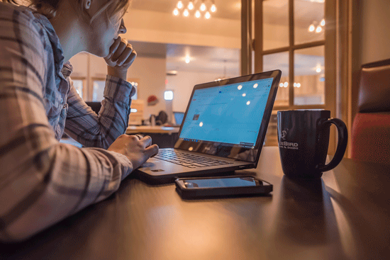 A photo of a woman wearing a flannel shirt sitting at a table looks at a laptop with her head in her hand and a phone and a black mug beside her.