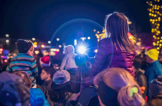 A photo of children sitting on adults' shoulders at a nighttime outdoor event with out of focus string lights in the background.
