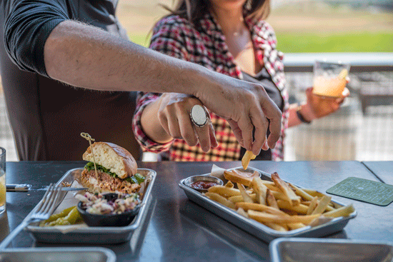 A photo of a woman and man, him wearing a dark shirt and her wearing a red flannel shirt and a large white ring on her index finger, both reaching for a plate of fries, their hands crossing each other.