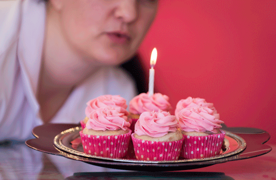A photo of a display of six cupcakes with pink icing and in pink wrappers on a pink background with a woman appearing to blow out a lit birthday candle placed in the middle of the display.