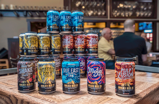 A photo of a display of Boulder Beer Company cans, from left to right showcasing Shake, Buffalo Gold, Due East, Mojo, and Pulp Fusion, arranged on a wooden table in a bar.