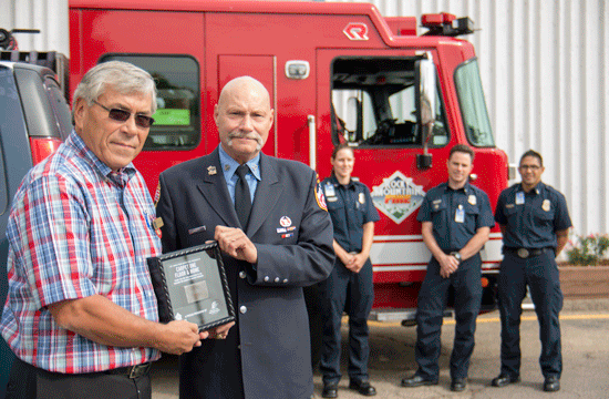 A man in a short-sleeve flannel shirt holds up a plaque with a man in official high-ranking uniform standing beside him, and three people standing in front of a red emergency services truck.