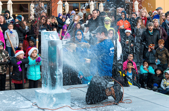 A photo of a man wearing a blue jacket and a baseball hat kneeling down in front of a clock of ice and sculpting it with a power tool watched by a crowd of onlookers, comprising people of all ages including children wearing Santa hats.