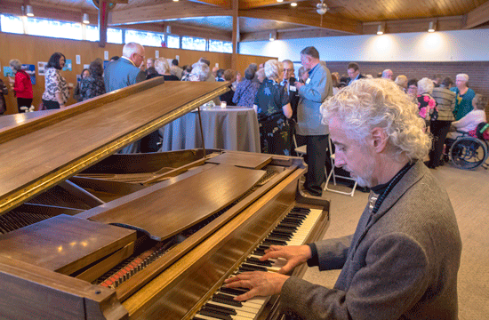 A photo of a man with white hair and a beard wearing a tweed blazer playing the piano for a conference room full of people and tables.