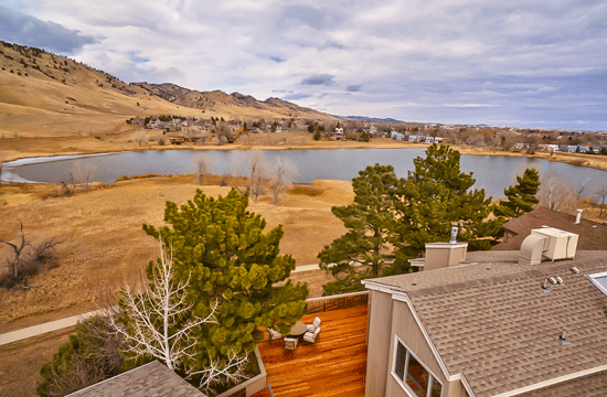 An aerial photo of the grey roof and wooden deck of a house, on which a lawn table and chairs can be seen, with some trees surrounding it and a lake and mountain range in the background.