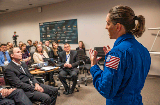 A photo of a woman with a blonde ponytail wearing a blue flightsuit with an american flag patch stitched onto the left arm giving a talk to a group of people sitting down seemingly looking at something behind her.