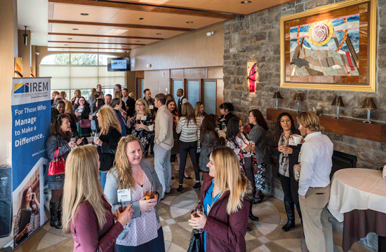A photo of attendees mingling while holding drinks at a conference with a banner reading "IREM, For Those Who Manage To Make A Difference."