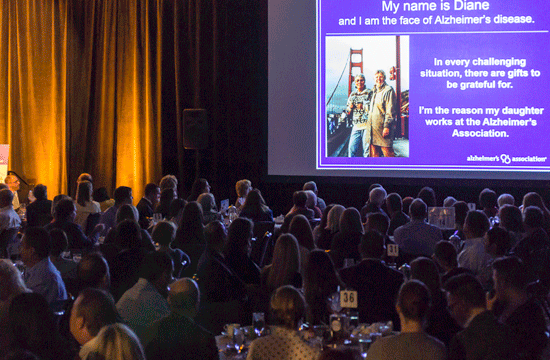 A photo of a roomful of people in a darkened hall seated at numbered tables look towards a screen with a photo of two people standing in front of the Golden Gate Bridge in San Francisco with text on a purple background titled "My Name is Diane and I am the Face of Alzheimer's Disease."