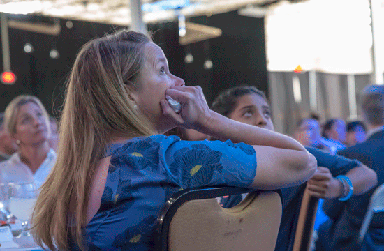 A photo of a woman with long blonde straight hair and a short-sleeved blue top or dress with flower motifs looking upwards with her chin in her hand in a conference space, while seated at a table surrounded by other people.