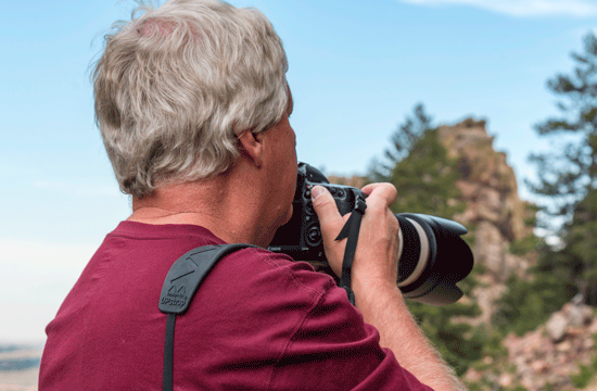 A photo of a man with grey hair and a maroon red shirt looks above a DLSR camera at a mountain area.