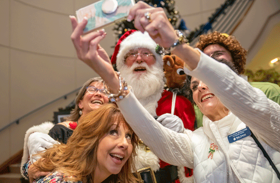 A photo of four people taking a selfie with a man dressed like Santa Claus, indoors, with a Christmas tree in the background.