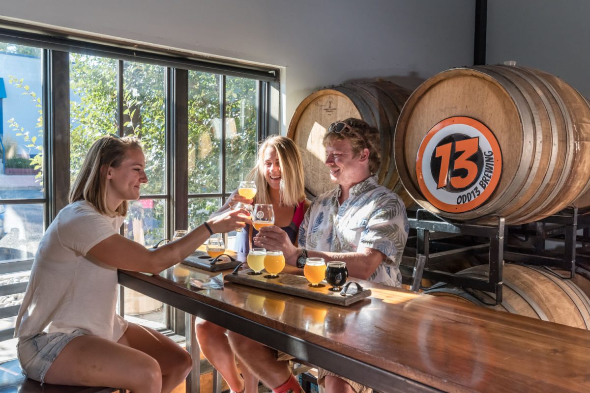 A photo of three people sit at a high bar detached bar area drinking a sample selection of craft beer and cheers their glasses while smiling, with a large window to their side and beer barrels behind them.