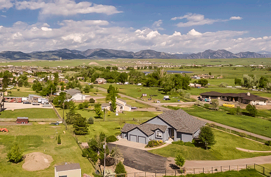 An aerial photo of a neighborhood surrounded by nature, fields, and trees, with a blue-walled house with grey roof in the foreground and a mountain range in the background.
