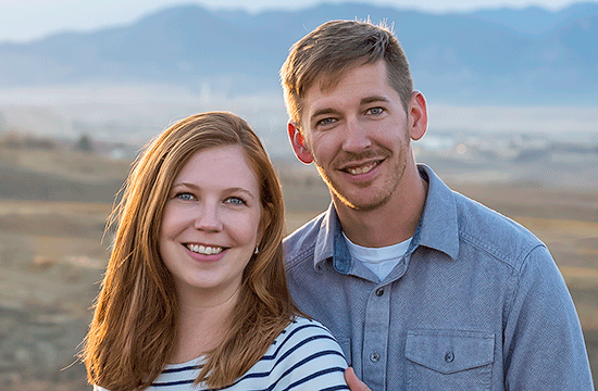 A photo of a young man and a woman standing in front of a mountain range smiling, the woman wearing a striped top and the man wearing a light blue button-up shirt and a white undershirt.