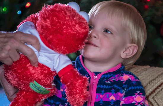 A photo of a young blonde child wearing a blue and pink sweater while an adult holds an Elmo stuffed animal up to their face from offscreen.
