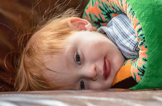 A photo of a red-headed young child looking at the camera wearing a striped shirt, lying sideways wrapped in a green blanket.