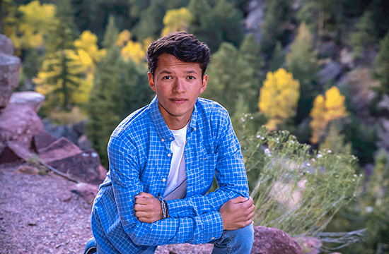 A photo of a young man with black hair kneeling outside in front of a forest, posing while wearing a white undershirt, a blue button-up shirt, jeans.