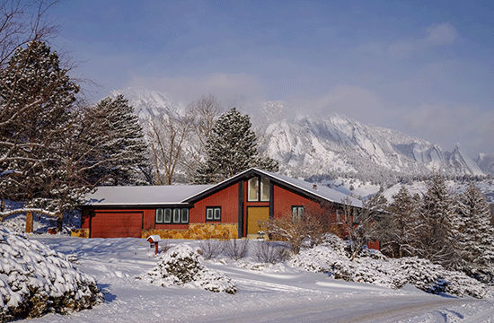 A photo of a wooden-looking house in a snowy landscape, with snow-capped mountains in the background.